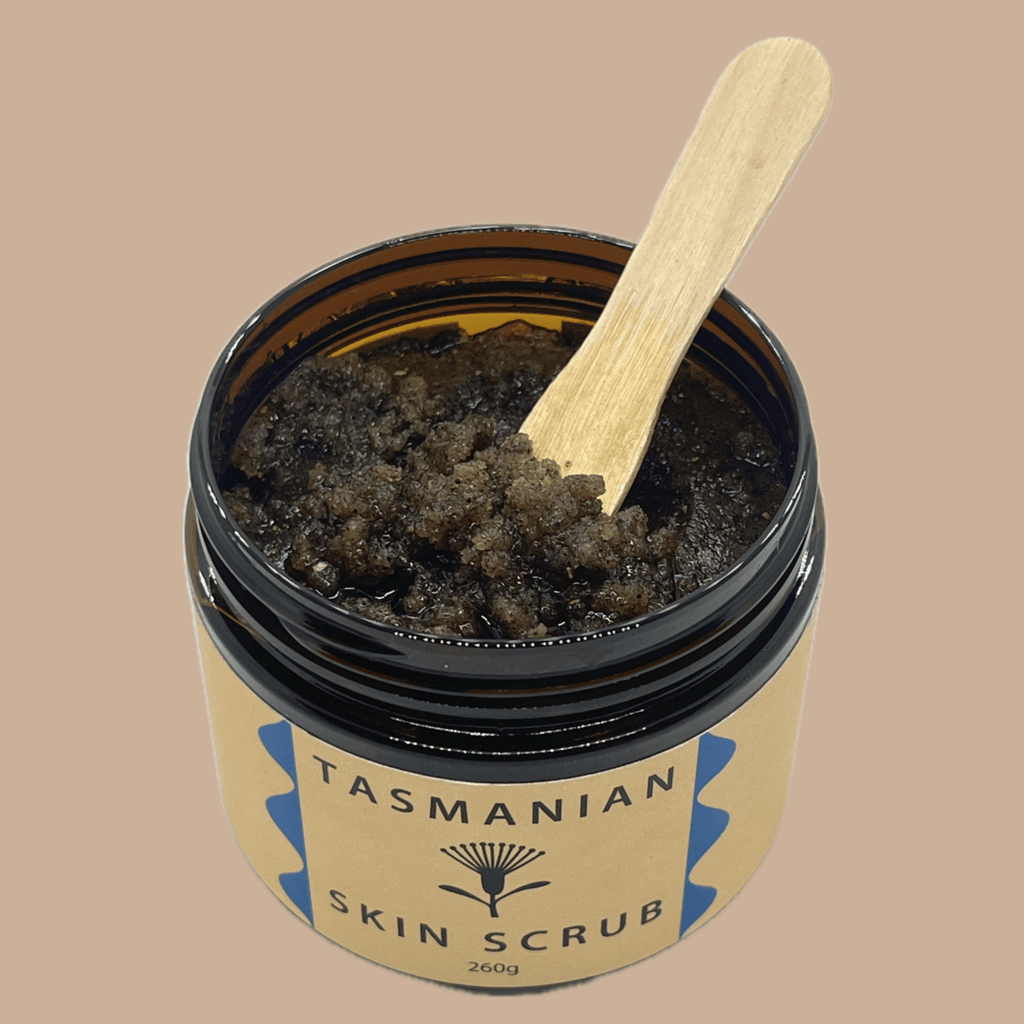 An open amber PET jar of Tasmanian Skin Scrub with a light wooden spatula resting inside, set against a solid beige background.