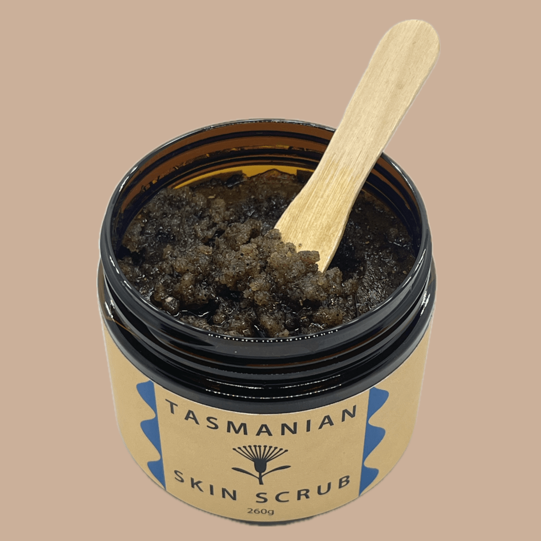 An open amber PET jar of Tasmanian Skin Scrub with a light wooden spatula resting inside, set against a solid beige background.
