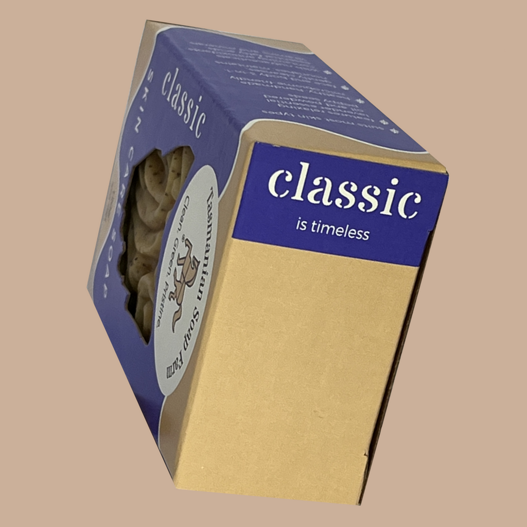 A boxed classic skin care soap, by Tasmanian Soap Farm, shown at an angle on a plain beige background. The package is primarily purple and kraft beige, with a visible oval cut out window showing a baroque-patterned soap inside.