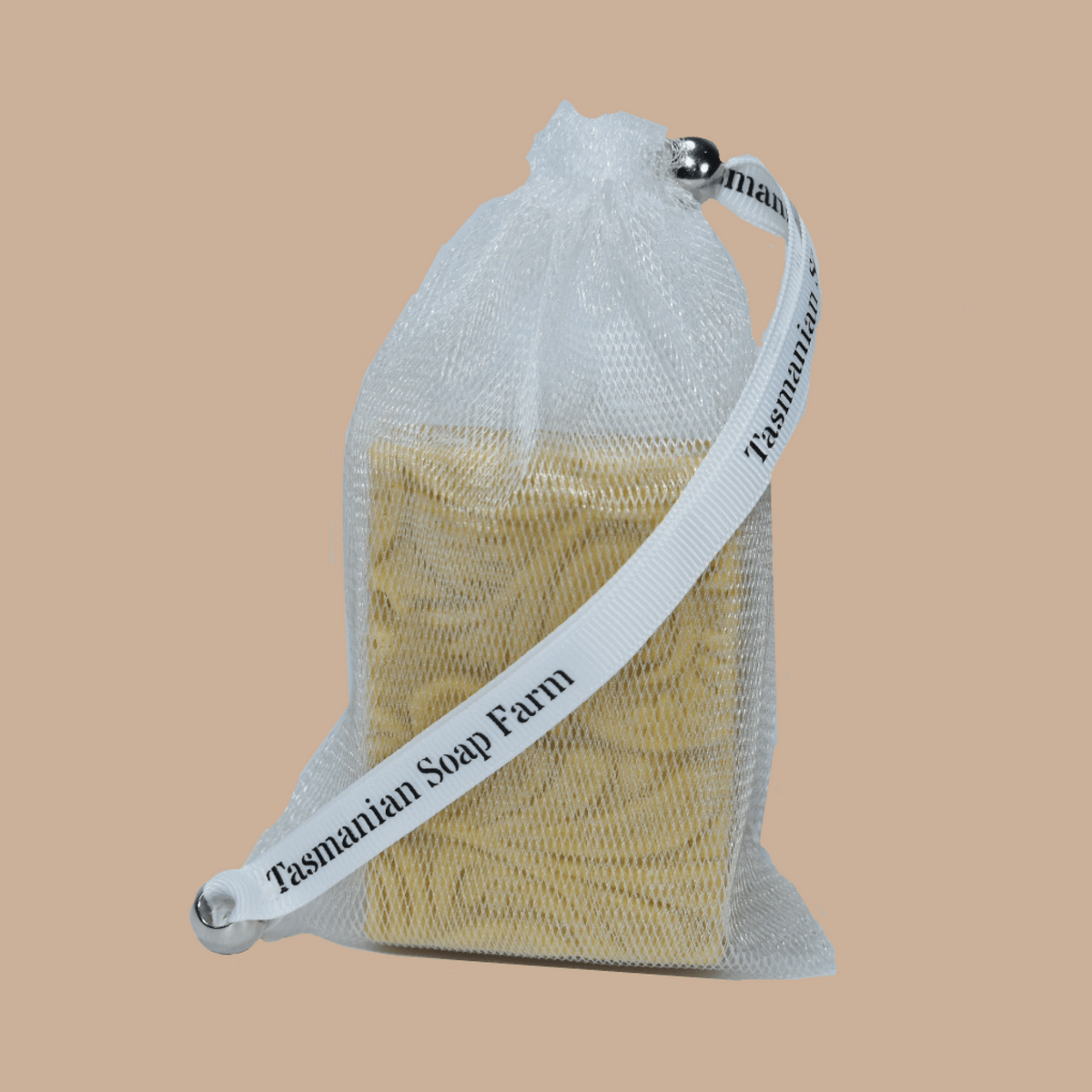 A white mesh drawstring pouch containing a bar of soap, with a white ribbon strap printed with the brand name 'Tasmanian Soap Farm', set against a solid beige background.