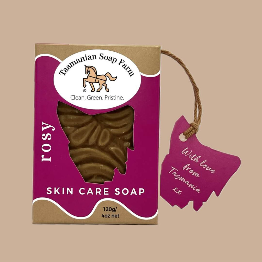 A package of Tasmanian Soap Farm Rosy Skin Care Soap, featuring a pink-hued soap bar visible through a window in a kraft paper box with a hot pink label, alongside a matching pink hang tag on a solid beige background.