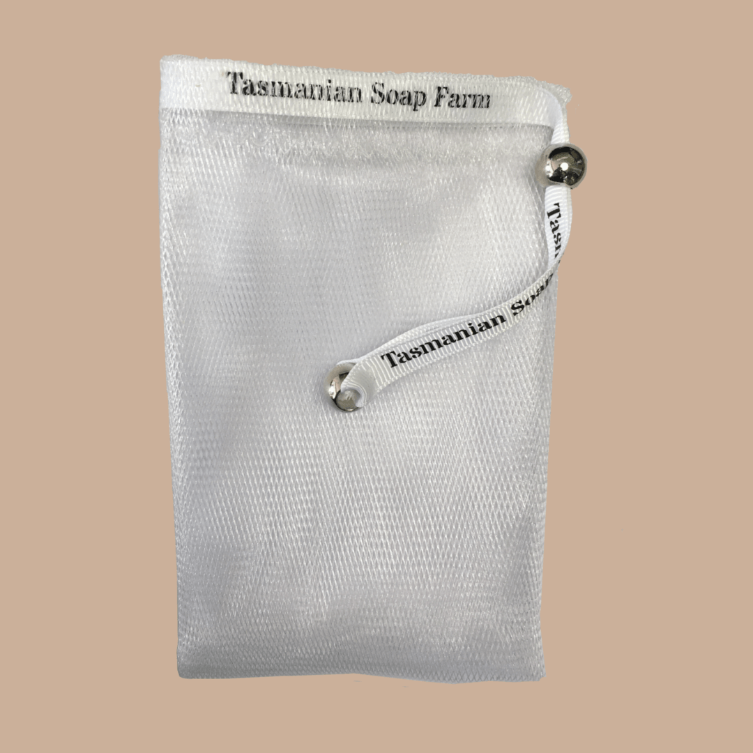 A white mesh soap bag with a white ribbon printed with the brand name 'Tasmanian Soap Farm' and two silver metal pony beads, shown against a solid beige background.