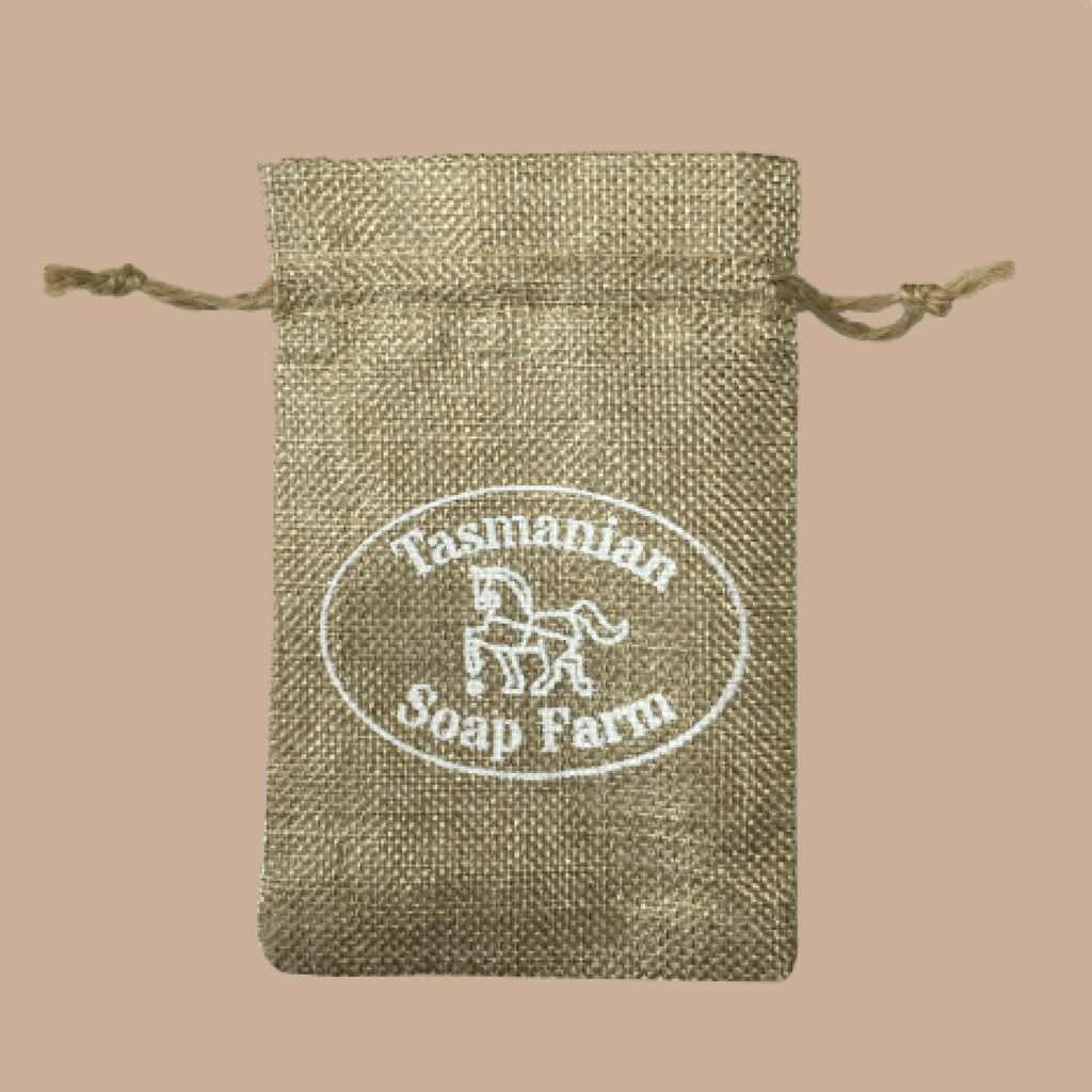A small natural jute drawstring pouch with a white printed oval logo reading 'Tasmanian Soap Farm' on a solid beige background.