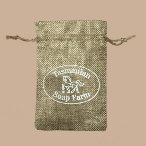 A small natural jute drawstring pouch with a white printed oval logo reading 'Tasmanian Soap Farm' on a solid beige background.