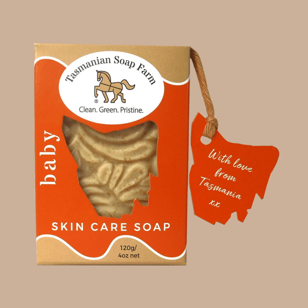 Tasmanian Soap Farm 'baby' skin care soap in a kraft paper box with an orange front panel, a clear window showing the pale beige soap bar, and a matching orange hangtag on a plain beige background.