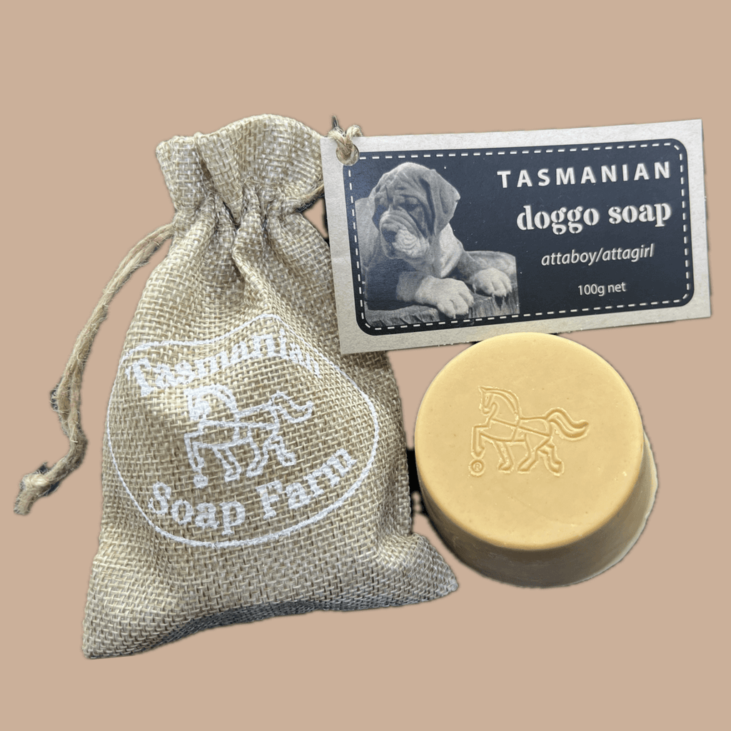 A 100g bar of Tasmanian doggo soap by Attaboy/Attagirl from Tasmanian Soap Farm, shown with its beige burlap drawstring pouch and a product tag featuring a puppy photo, set against a solid tan background.