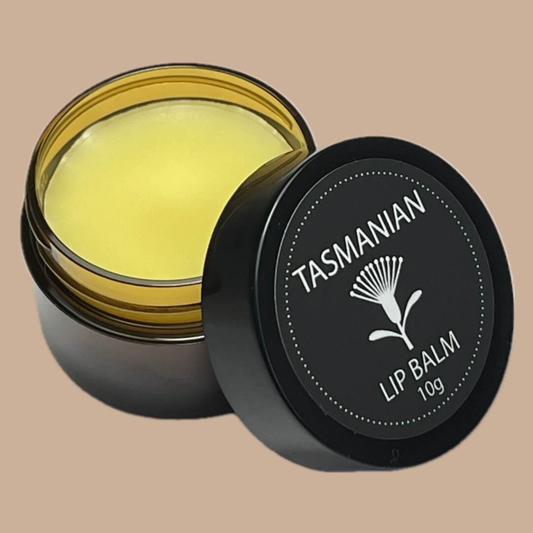 An open amber PET pot of Tasmanian lip balm showing the pale yellow balm inside, with its black lid leaning against it. The lid features a white floral logo and the text 'TASMANIAN LIP BALM 10g'.