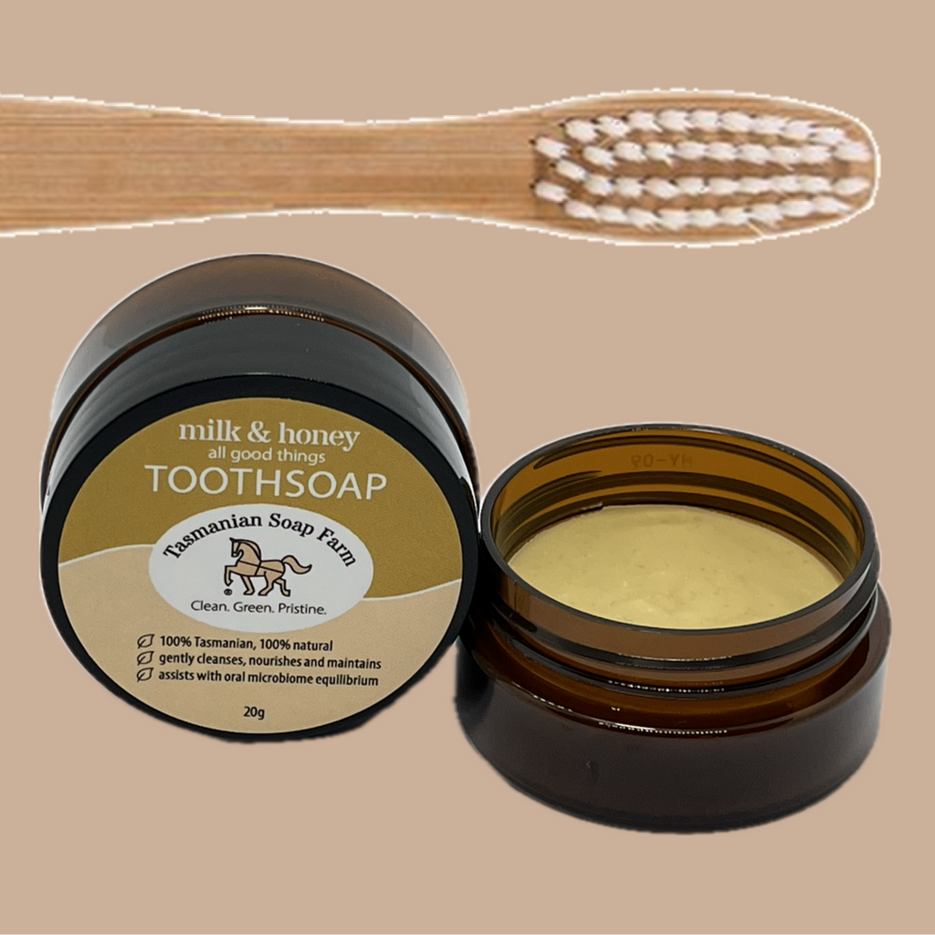 An open pot of milk & honey Tasmanian Soap Farm toothsoap shown next to its closed one, with a wooden toothbrush at the top on a solid beige background.
