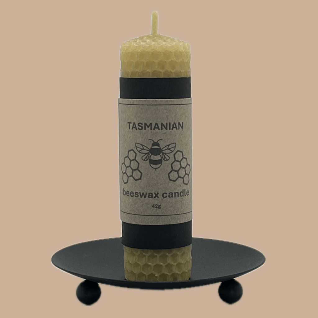 A Tasmanian beeswax candle in a natural yellow-green hue, displayed on a black metal candle holder against a solid beige background. The candle features a paper label with a bee and honeycomb design.