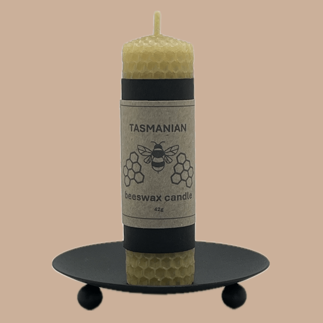 A Tasmanian beeswax candle in a natural yellow-green hue, displayed on a black metal candle holder against a solid beige background. The candle features a paper label with a bee and honeycomb design.