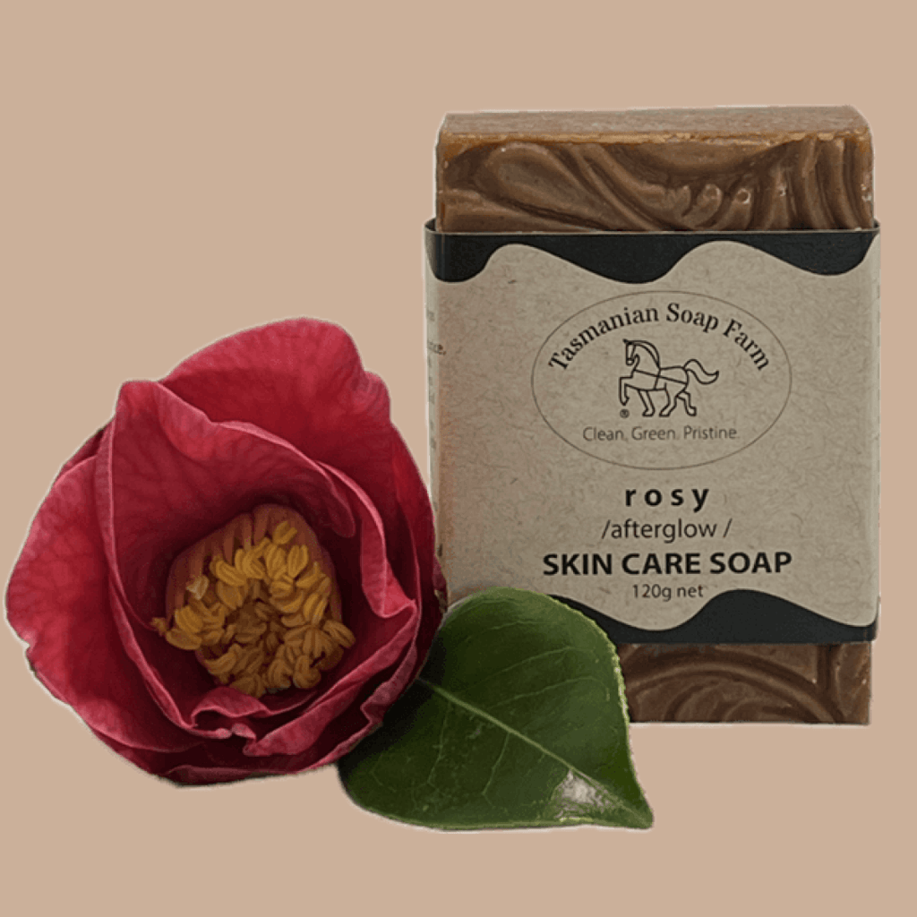 A bar of Tasmanian Soap Farm 'rosy /afterglow /' skin care soap, wrapped in a kraft paper label with a black horse logo, shown against a solid beige background with a pink rose graphic overlay in the lower right corner.