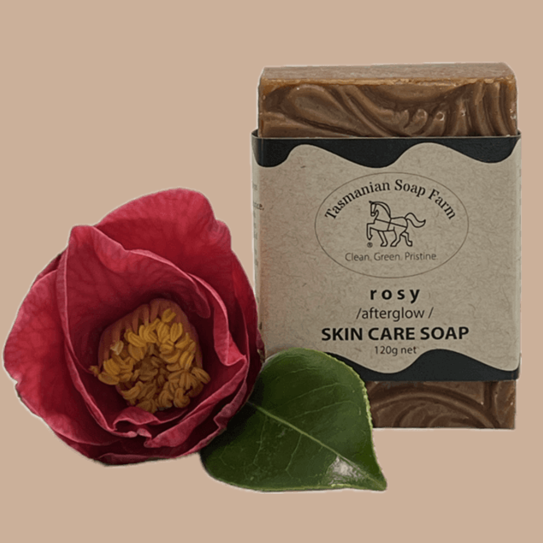 A bar of Tasmanian Soap Farm 'rosy /afterglow /' skin care soap, wrapped in a kraft paper label with a black horse logo, shown against a solid beige background with a pink rose graphic overlay in the lower right corner.