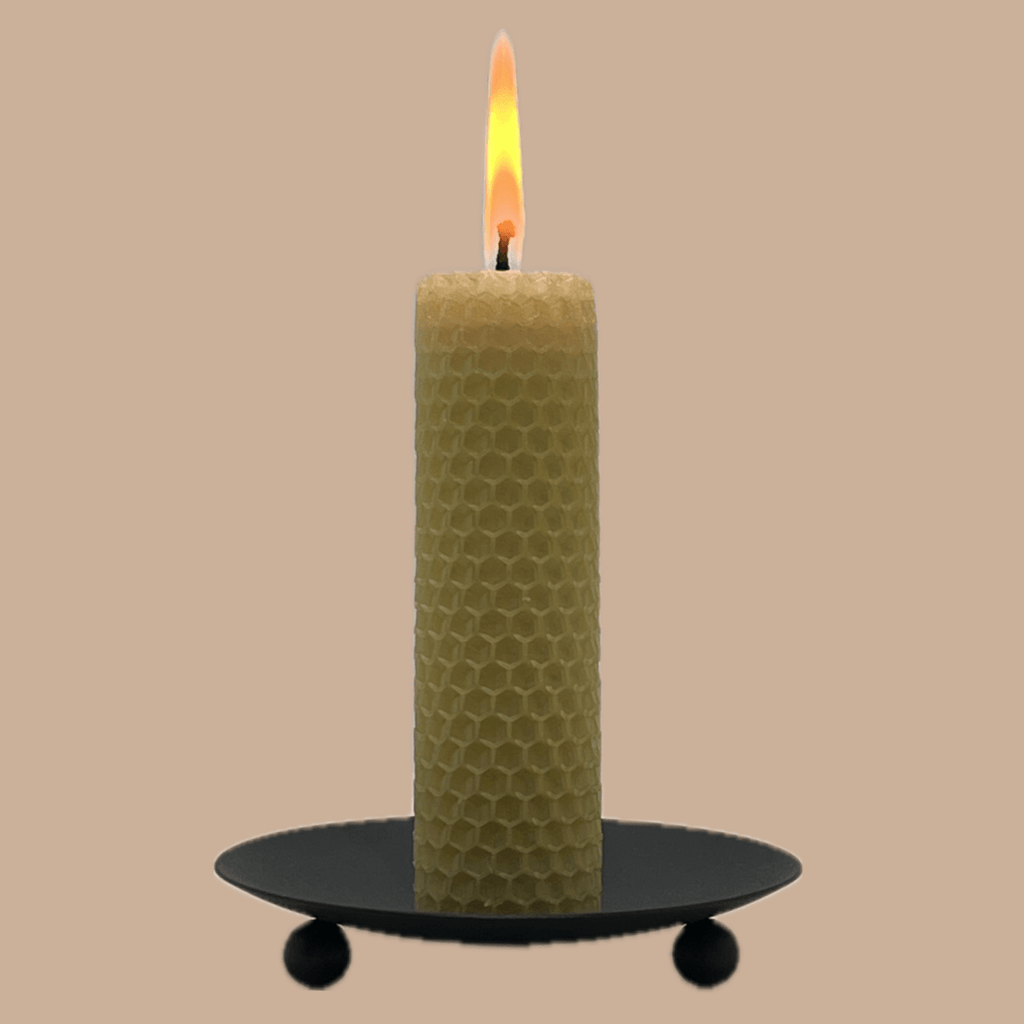A lit honeycomb-patterned candle in a pale yellow-green shade, standing on a black metal candle holder with three small feet against a solid beige background.