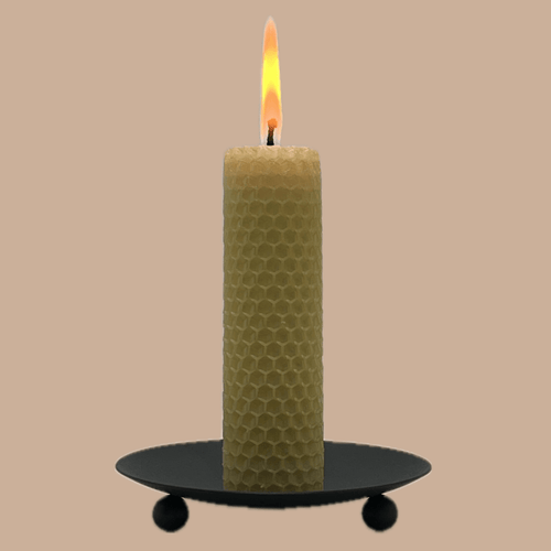 A lit honeycomb-patterned candle in a pale yellow-green shade, standing on a black metal candle holder with three small feet against a solid beige background.