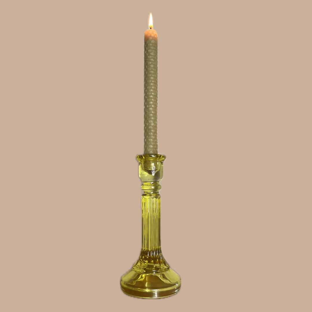 A lit taper candle with a ribbed beige-wrapped upper section and a translucent yellow glass holder, centered against a solid beige background.