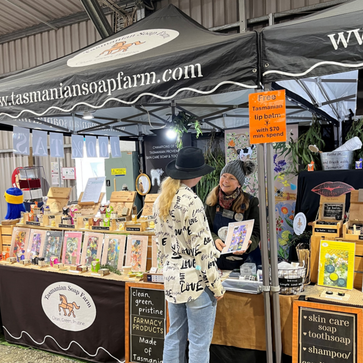 A market booth for Tasmanian Soap Farm under a black branded canopy tent, with a woman in a black hat and patterned shirt talking to the founder, Michelle Bennett, inside. The booth displays handmade soaps, bath products, and other artisan items.