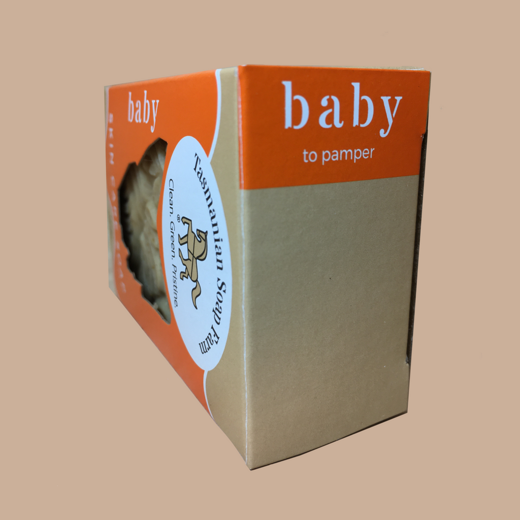 A boxed baby skin care soap, by Tasmanian Soap Farm, shown at an angle on a solid beige background. The packaging features a bright orange top and side panel with white text reading 'baby, to pamper', a kraft brown lower section, and a white oval sticker that says 'Tasmanian Soap Farm, clean, green, pristine'.