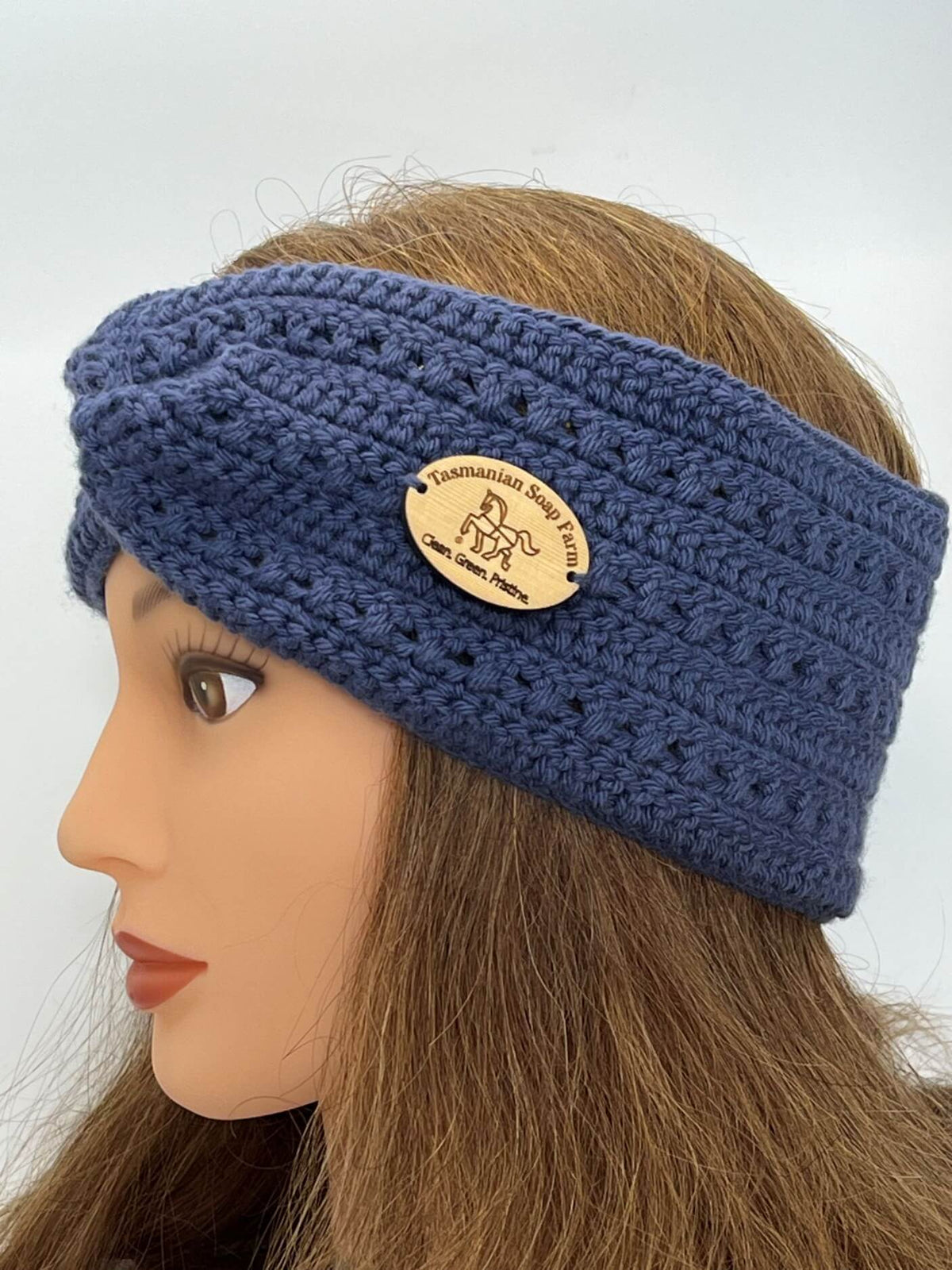 A French blue crocheted headband with a wooden brand plaque, displayed on a mannequin head with long light brown hair against a plain light grey background.