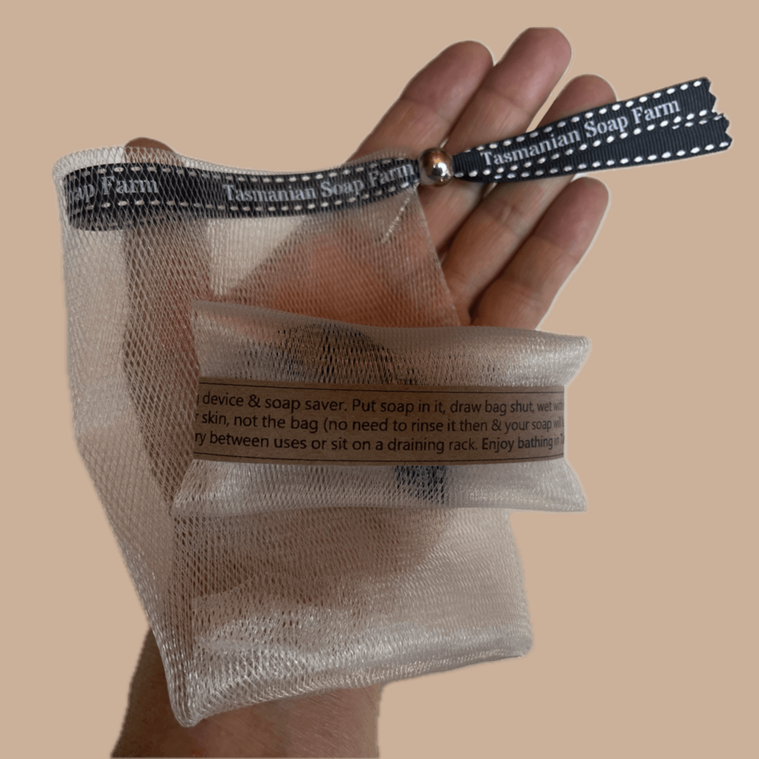 A hand holding a Tasmanian Soap Farm reusable mesh soap saver bag with a black branded strap, shown against a solid beige background.