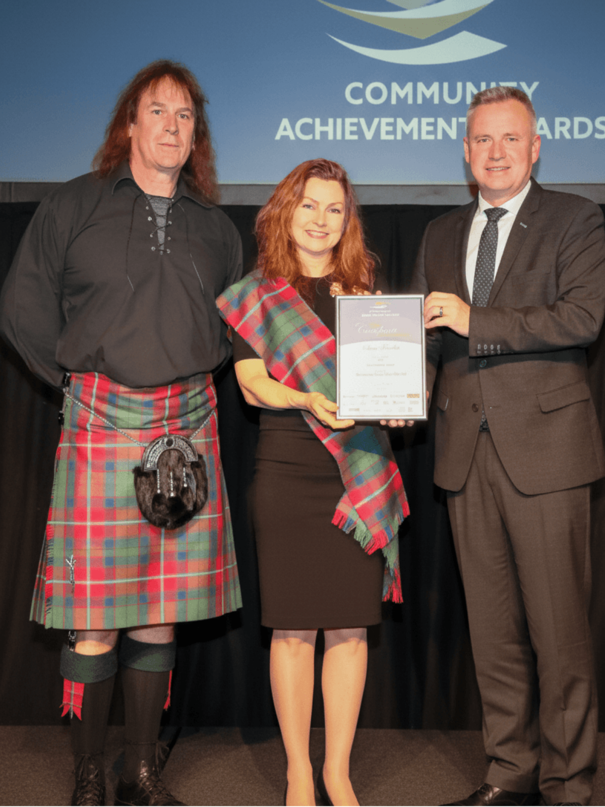Nick and Michelle Bennett of Tasmanian Soap farm are standing receiving an award from Premier Jeremy Rockliff at a Community Achievement Awards event. Nick wears a black shirt and a red tartan kilt with a sporran. Michelle is in a black dress with a matching tartan sash.