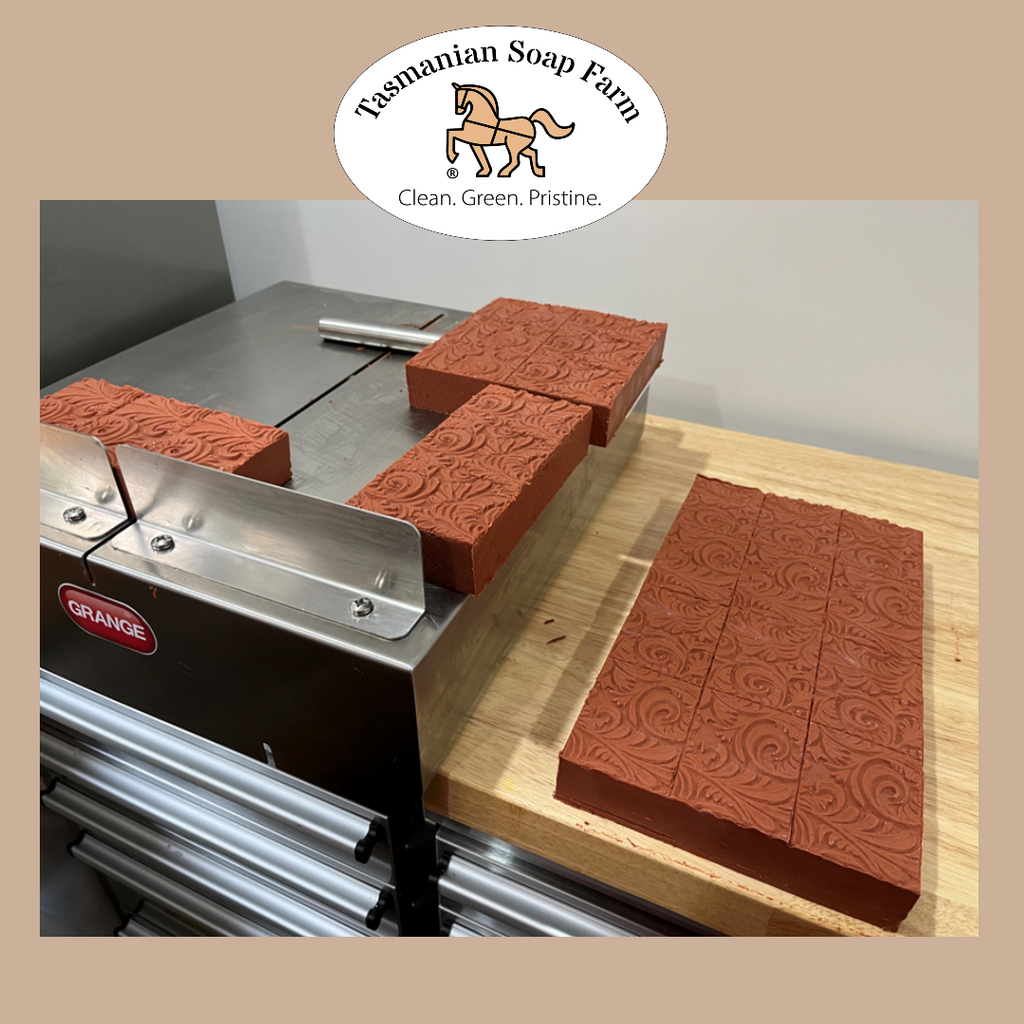 A slab of embossed terracotta-colored soap shown on a wooden table next to a stainless steel machine, with other soap slabs and an oval 'Tasmanian Soap Farm' logo overlay at the top.