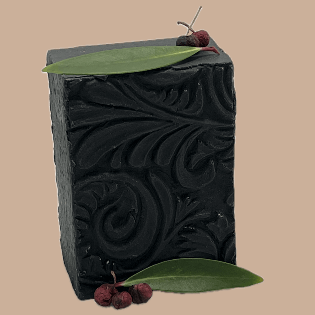 A rectangular bar of dark charcoal-coloured soap with embossed scrollwork, decorated with a green leaf and small dried red pepper berries, set against a solid beige background.
