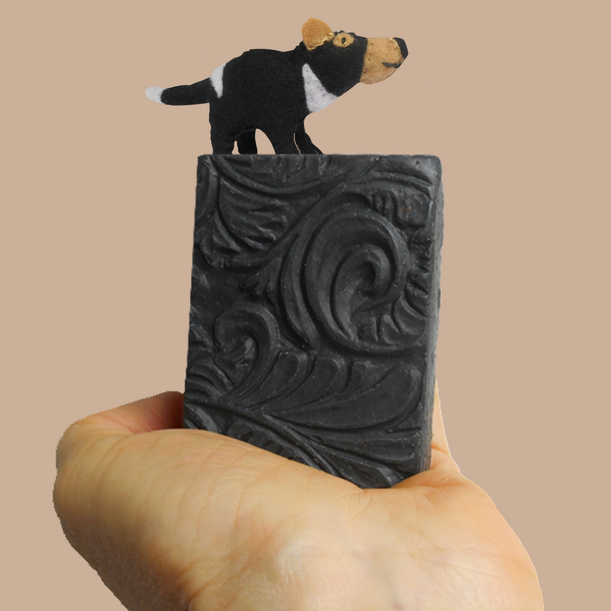 A hand holding a rectangular black soap bar with an embossed scroll pattern, topped with a small sculpted dog figurine in black, tan, and white, against a solid beige background. It's Tasmanian Soap Farm's devil skin care soap.