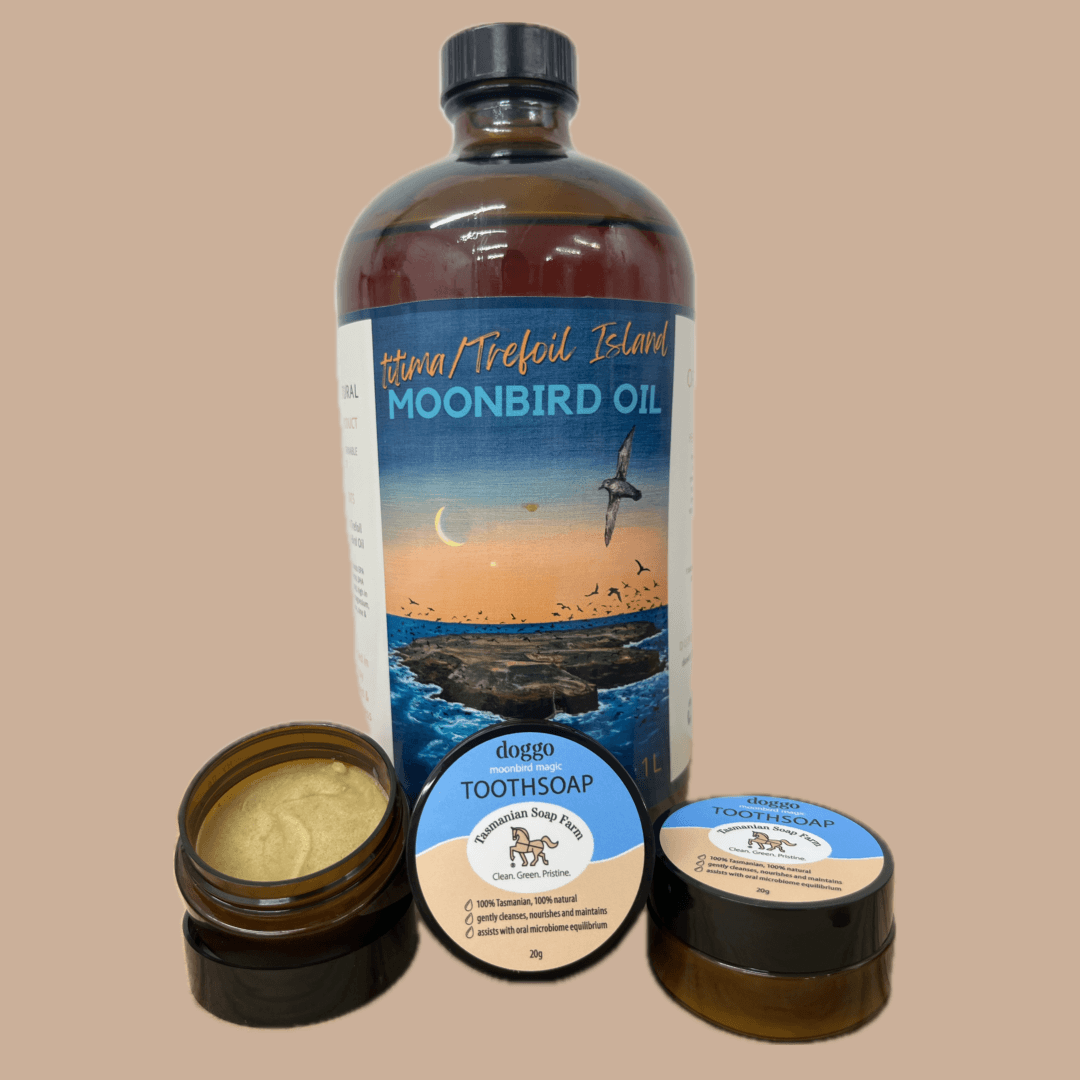 A bottle of  titima/Trefoil Island Moonbird Oil with two small pots of doggo toothsoap in front, one open showing a pale yellow cream and one closed with a blue and white label. The products are arranged on a solid beige background.