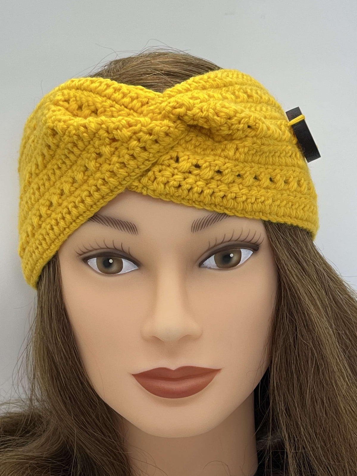 A yellow crocheted headband with a twisted front detail, shown on a mannequin head with long brown hair against a plain light grey background.