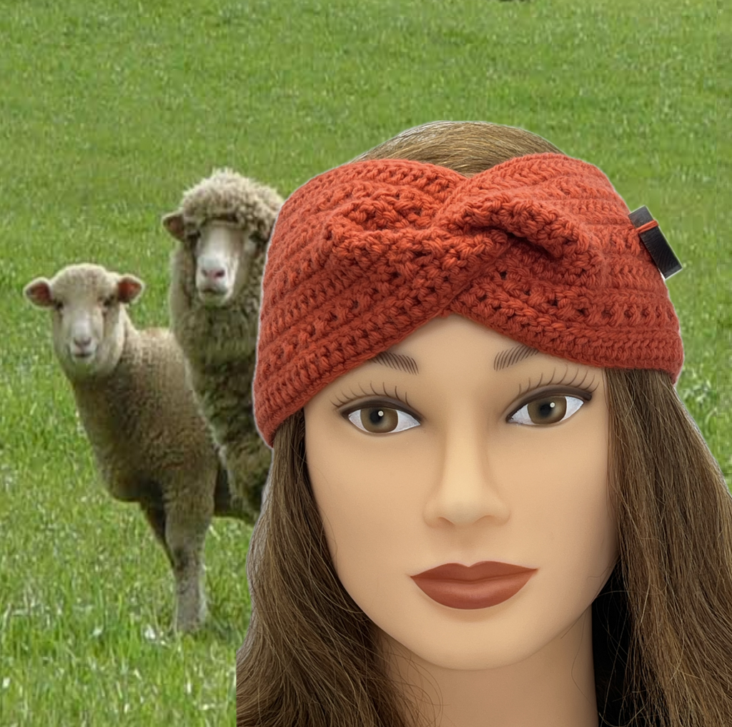 A close-up of a mannequin head wearing a textured twisted knit headband in coral, with a blurred background of two sheep on green grass.