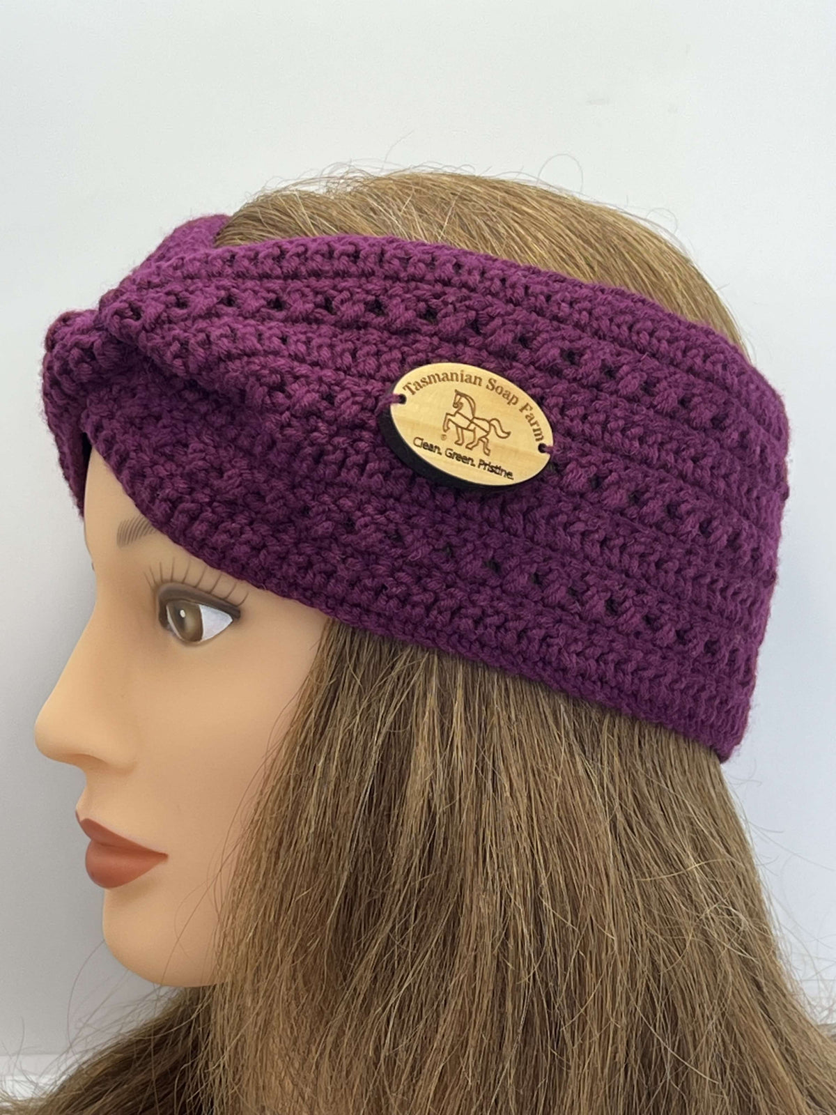 A purple crocheted twist-front ear warmer headband by Tasmanian Soap Farm with a round tan timber logo patch, displayed on a mannequin head with light brown hair against a plain white background.
