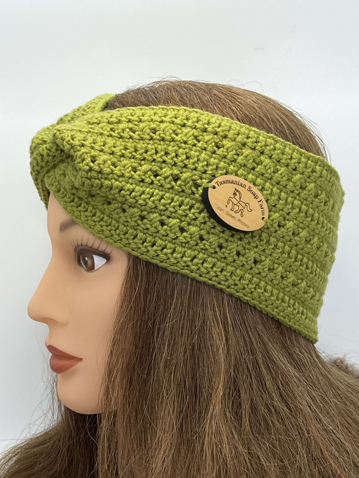 A close-up side profile of a mannequin head wearing a chunky grass green crocheted twist-front headband with a round wooden brand tag that reads 'Tasmanian Soap Farm'.
