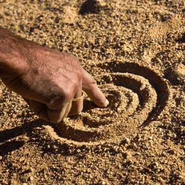 An Australian first nations person drawing a circular pattern in sand.