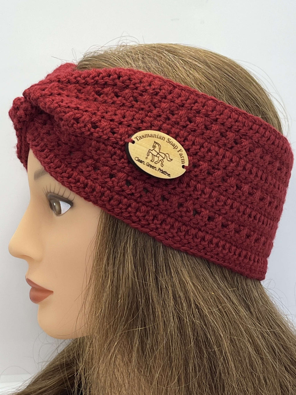 A close-up side view of a mannequin head wearing a chunky crocheted twist-front headband in deep burgundy red. The headband features a natural wood oval brand patch that reads 'Tasmanian Soap Farm'.