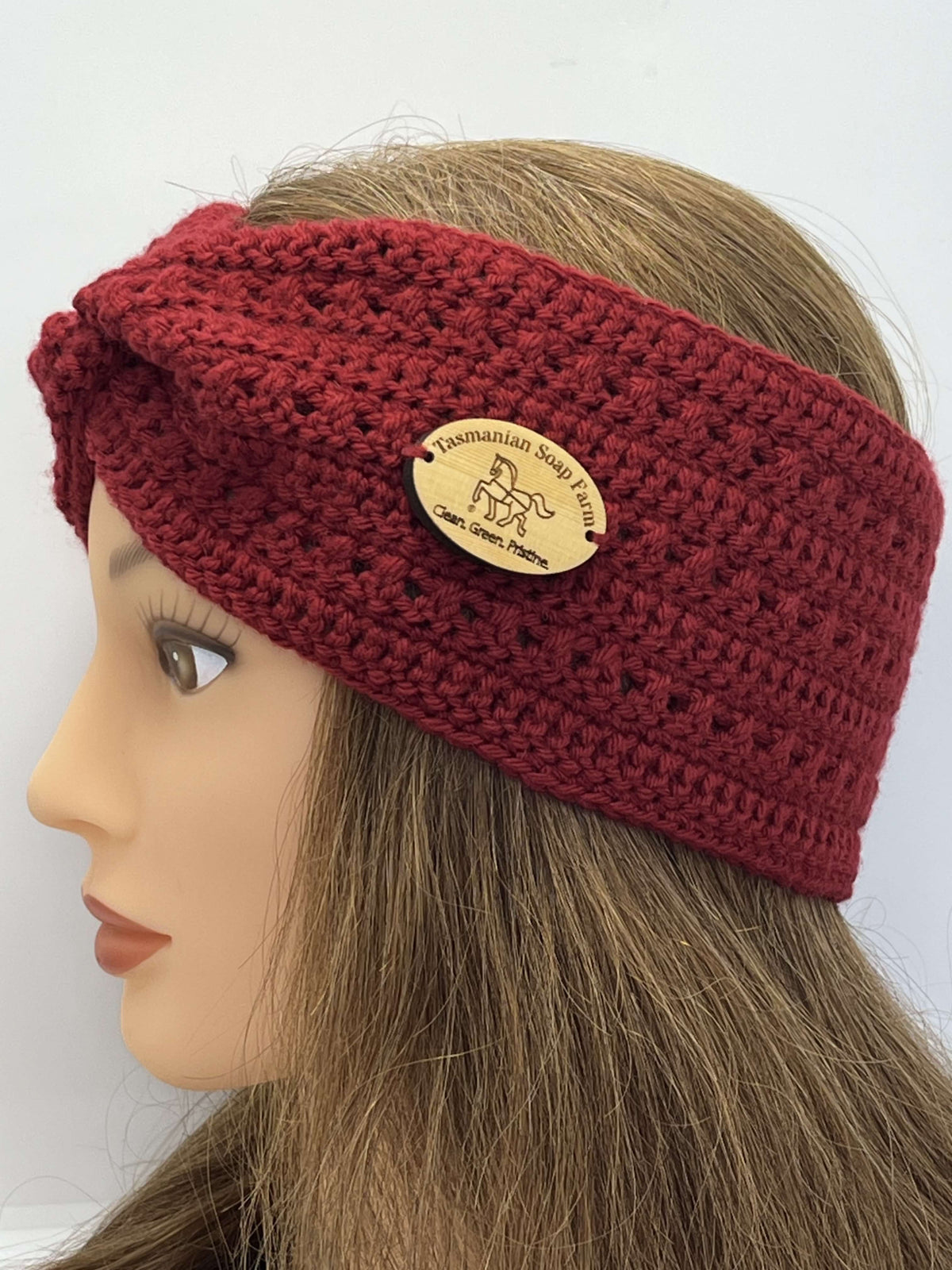 A close-up side view of a mannequin head wearing a chunky crocheted twist-front headband in deep burgundy red. The headband features a natural wood oval brand patch that reads 'Tasmanian Soap Farm'.