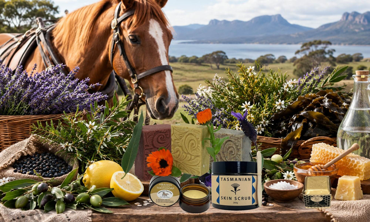 A styled outdoor display of Tasmanian Soap Farm products and ingredients featuring a horse in the background with lavender, flowers, lemons, honey, and local ingredients arranged around several skincare items, including a tub labelled Tasmanian Skin Scrub, on a rustic table with a scenic mountain and lake landscape behind.