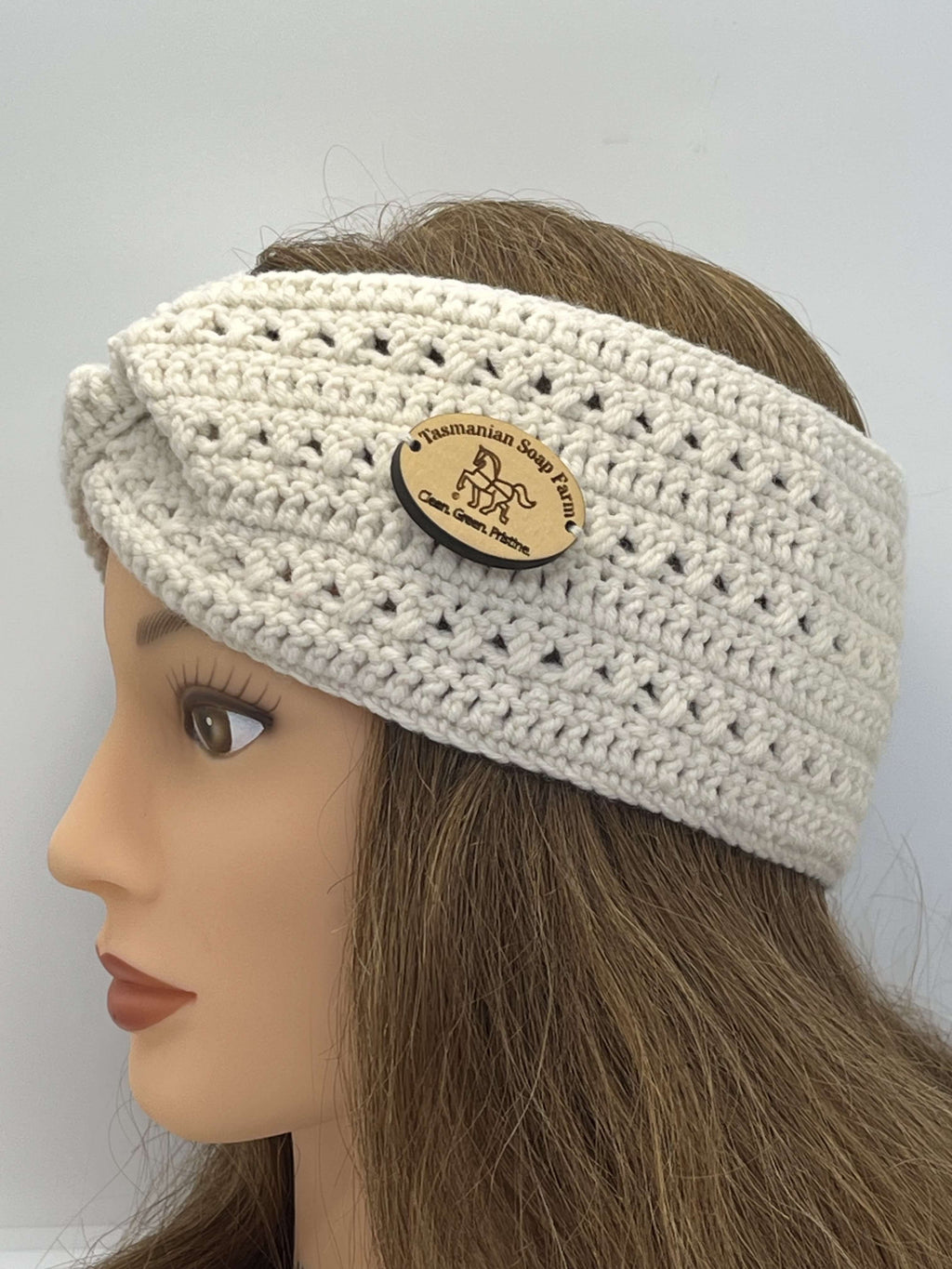 A cream-coloured crocheted headband with a textured openwork pattern and a round wooden brand badge, displayed on a mannequin head with brown hair against a plain light grey background.