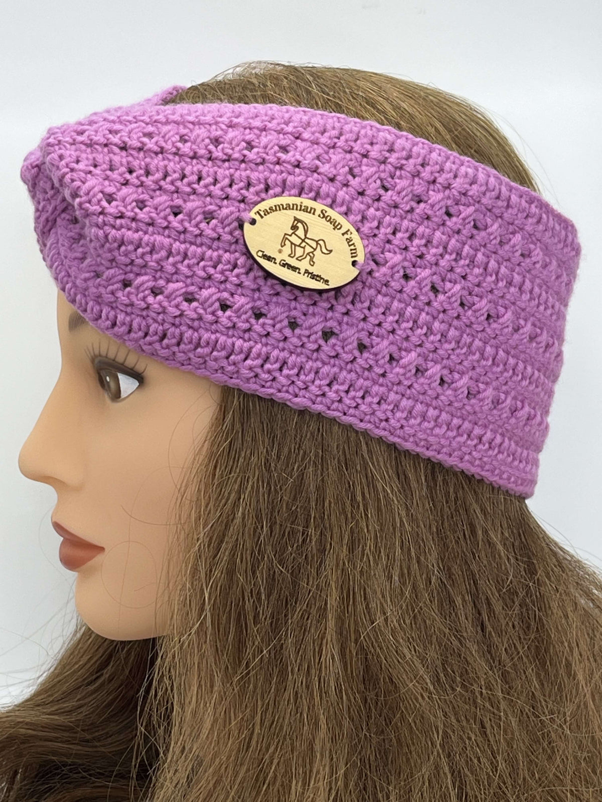 A pink crocheted headband by Tasmanian Soap Farm with a small oval wooden brand patch, shown on a mannequin head with long brown hair against a plain white background.