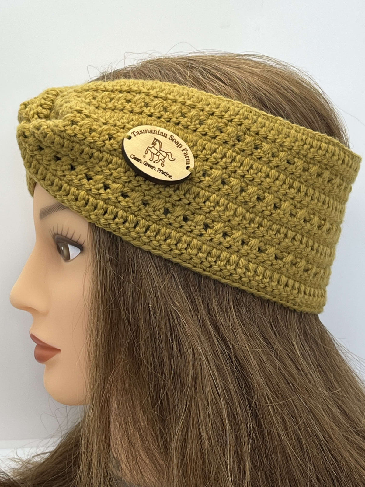 A mustard yellow Tasmanian Soap Farm crocheted headband with a wooden brand plaque, displayed on a mannequin head with long brown hair against a plain white background.