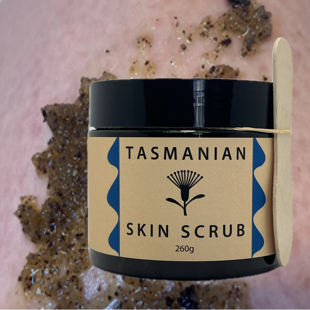 A tub of Tasmanian Skin Scrub with a kraft paper label and black lid, shown with a wooden spatula and a smear of the dark scrub on skin in the background.