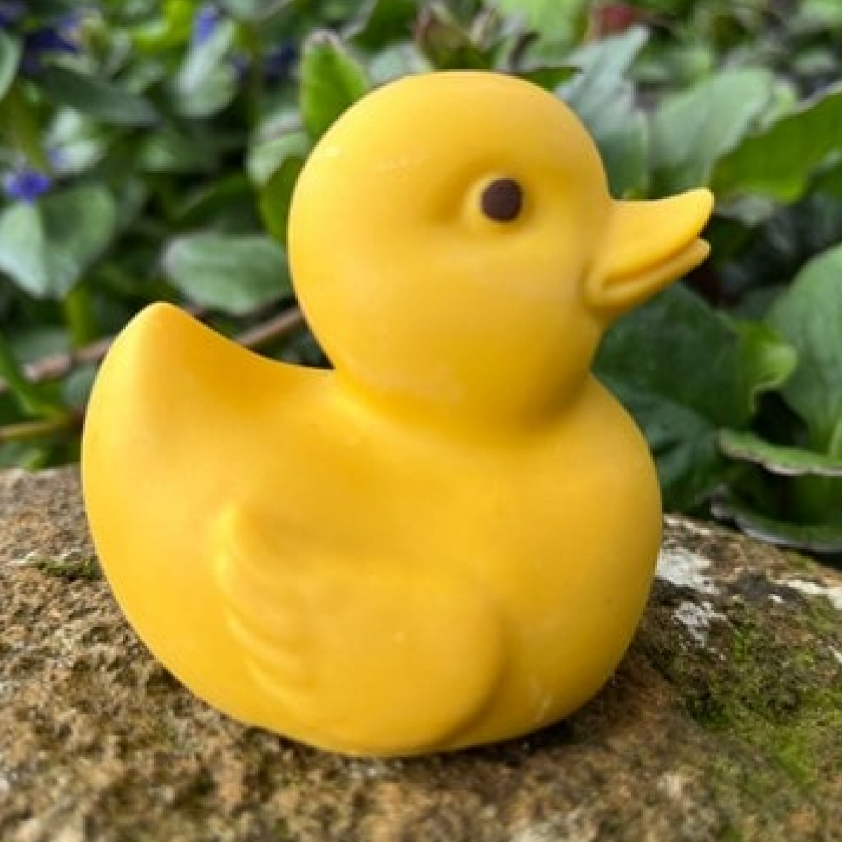 A yellow soap duck positioned on a mossy stone with a blurred background of green foliage.