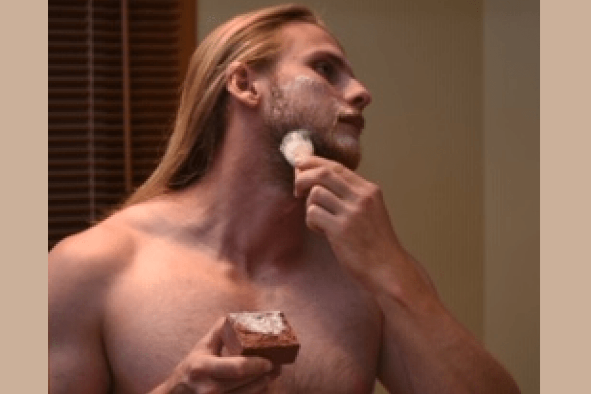 A shirtless man with long blond hair applies shaving soap to his face with a brush while holding a soap bar from Tasmanian Soap Farm in a bathroom setting.