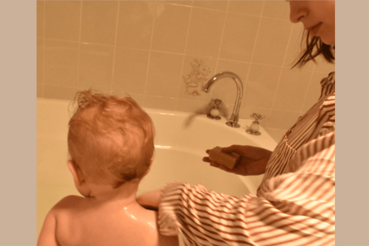 A baby with wet hair sits in a white bathtub while an adult in a striped shirt washes the baby's back with baby soap from Tasmanian Soap Farm in a tiled bathroom.