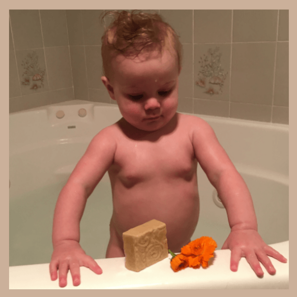 A baby standing in a bathtub holding a beige decorative Tasmanian Soap Farm baby soap bar and an orange flower, with a tiled bathroom background.