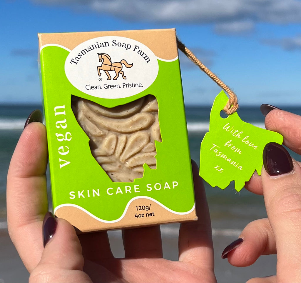 A person holds a box of vegan skin care soap from Tasmanian Soap Farm in front of a beach background on Bruny Island, Tasmania. The box is bright green and tan, with a clear window showing the soap inside, and a matching green hangtag is attached with a string.