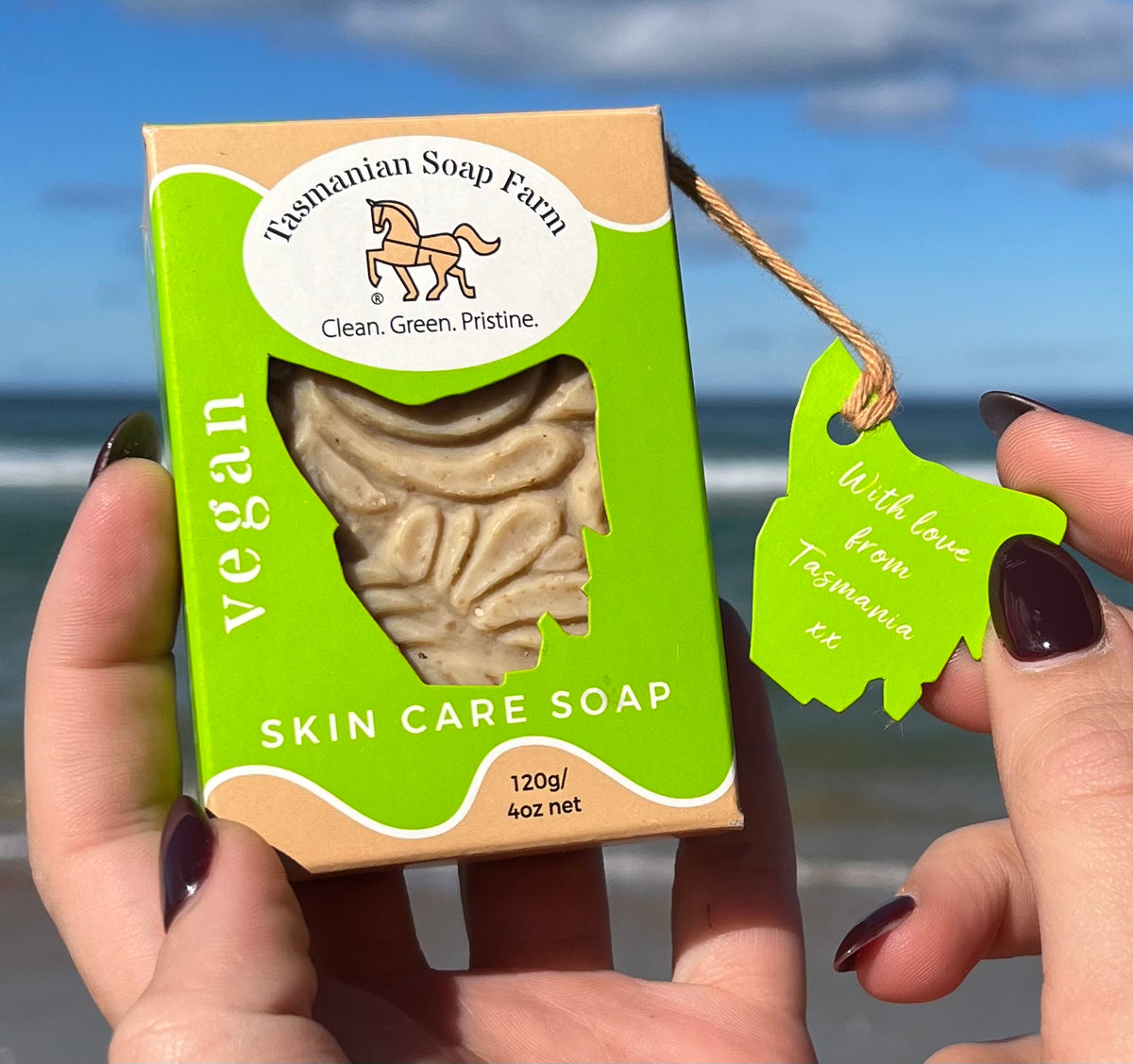 A person holds a box of vegan skin care soap from Tasmanian Soap Farm in front of a beach background on Bruny Island, Tasmania. The box is bright green and tan, with a clear window showing the soap inside, and a matching green hangtag is attached with a string.