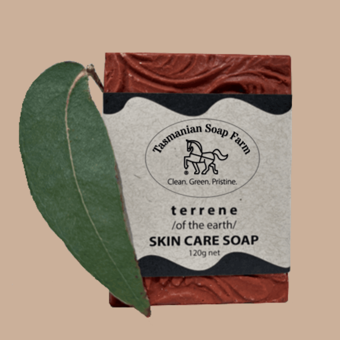 A bar of Tasmanian Soap Farm 'terrene' skin care soap with a textured terracotta red wrapper and a cream paper label, shown next to a green leaf on a solid beige background.