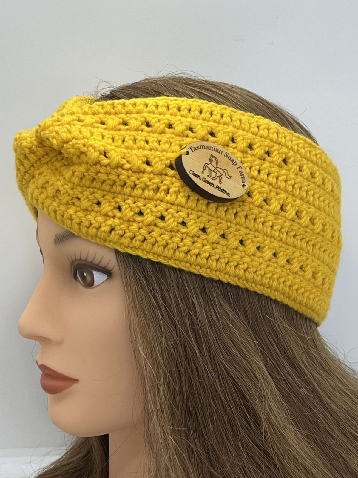 A bright yellow crocheted headband with a knotted side detail and a round wooden brand plaque, displayed on a mannequin head with brown hair against a plain light background.