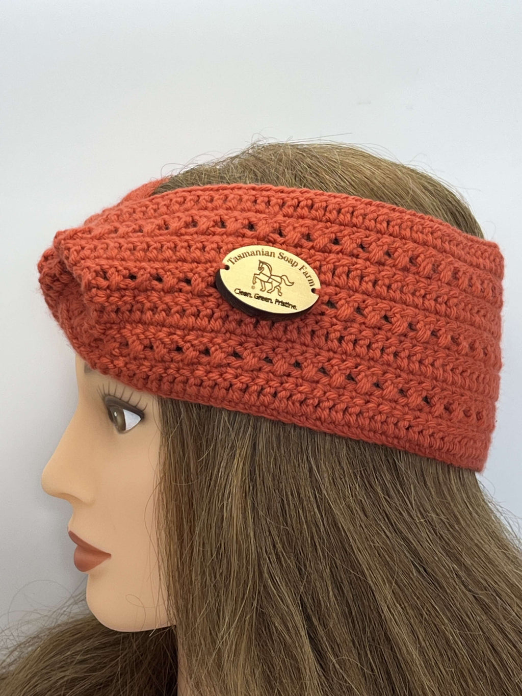 A coral crocheted headband with a small oval gold-tone logo charm, displayed on a mannequin head with long brown hair against a plain white background.