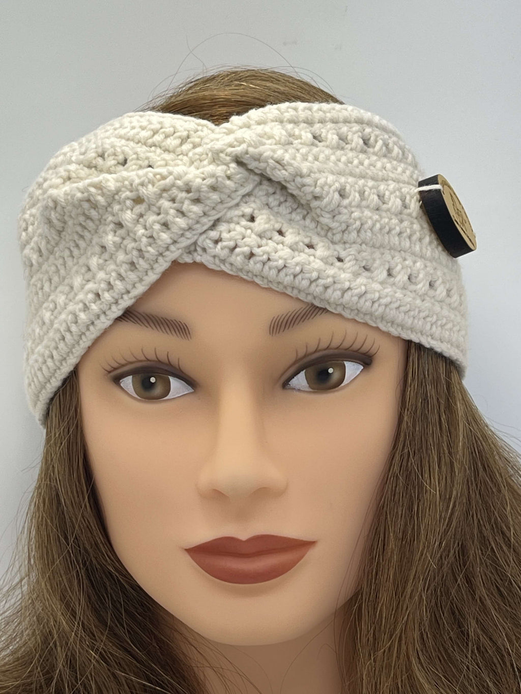 A cream-colored crocheted twist-front headband with a decorative timber button, displayed on a mannequin head with brown hair against a plain light grey background.